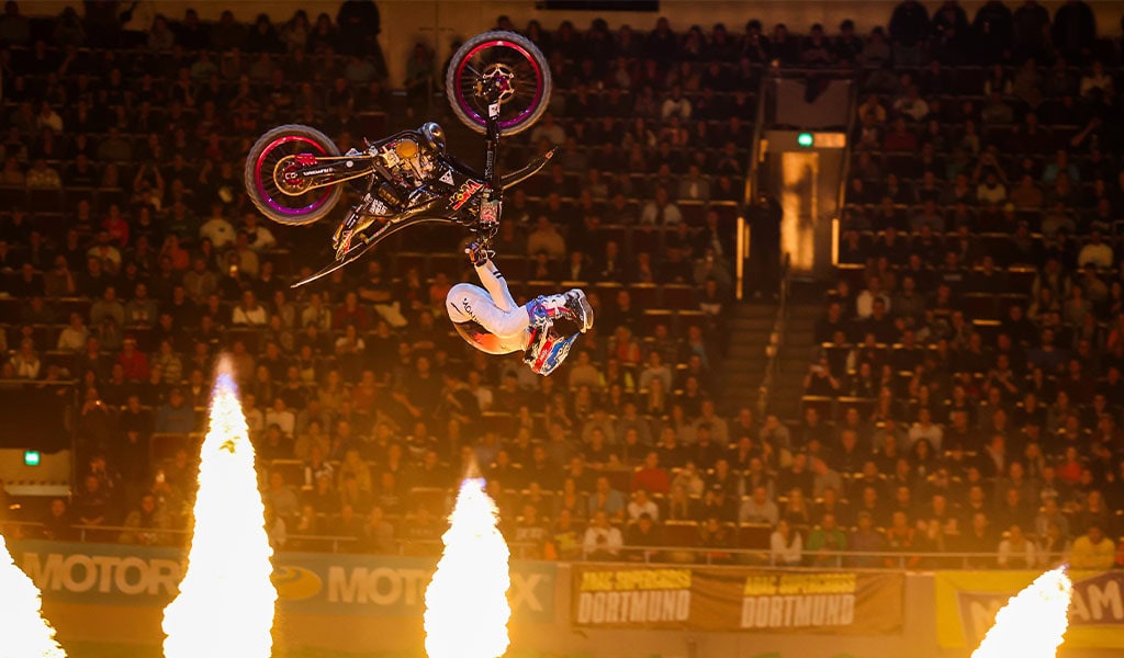 Motocross-Fahrer in der Luft, macht einen Backflip. Feuersäulen im Vordergrund. Publikum im Hintergrund.