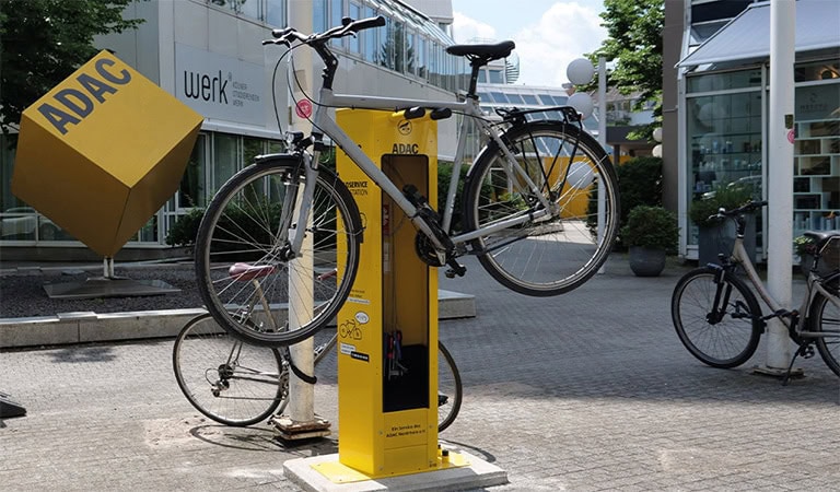 Eine gelbe ADAC-Radstation mit einem silbernen Fahrrad vorne, daneben Würfel-Skulptur und Gebäude.